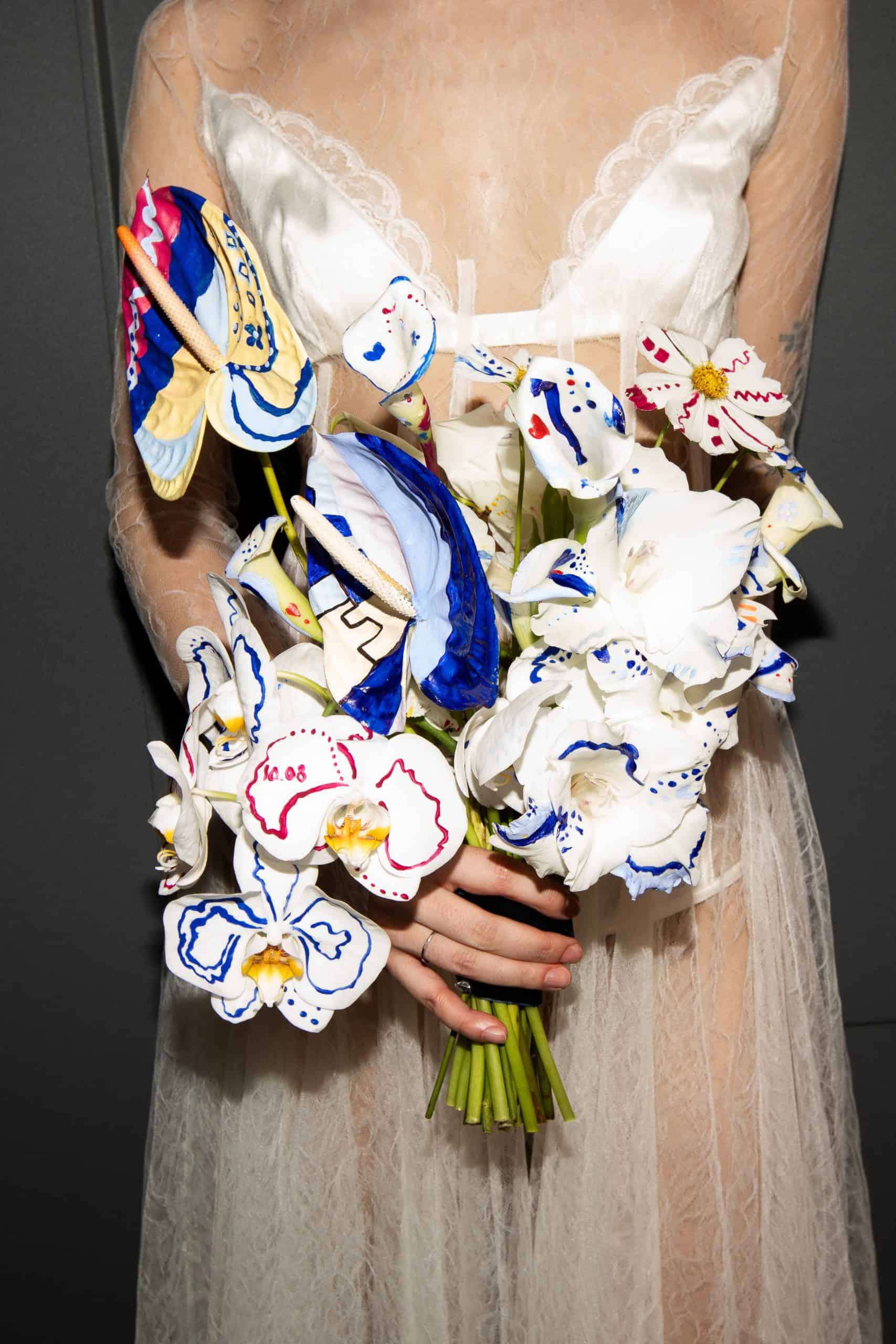 Hand-painted bridal bouquet held against a tulle dress, featuring sculptural flowers with vivid blue, red, and yellow brushstrokes.