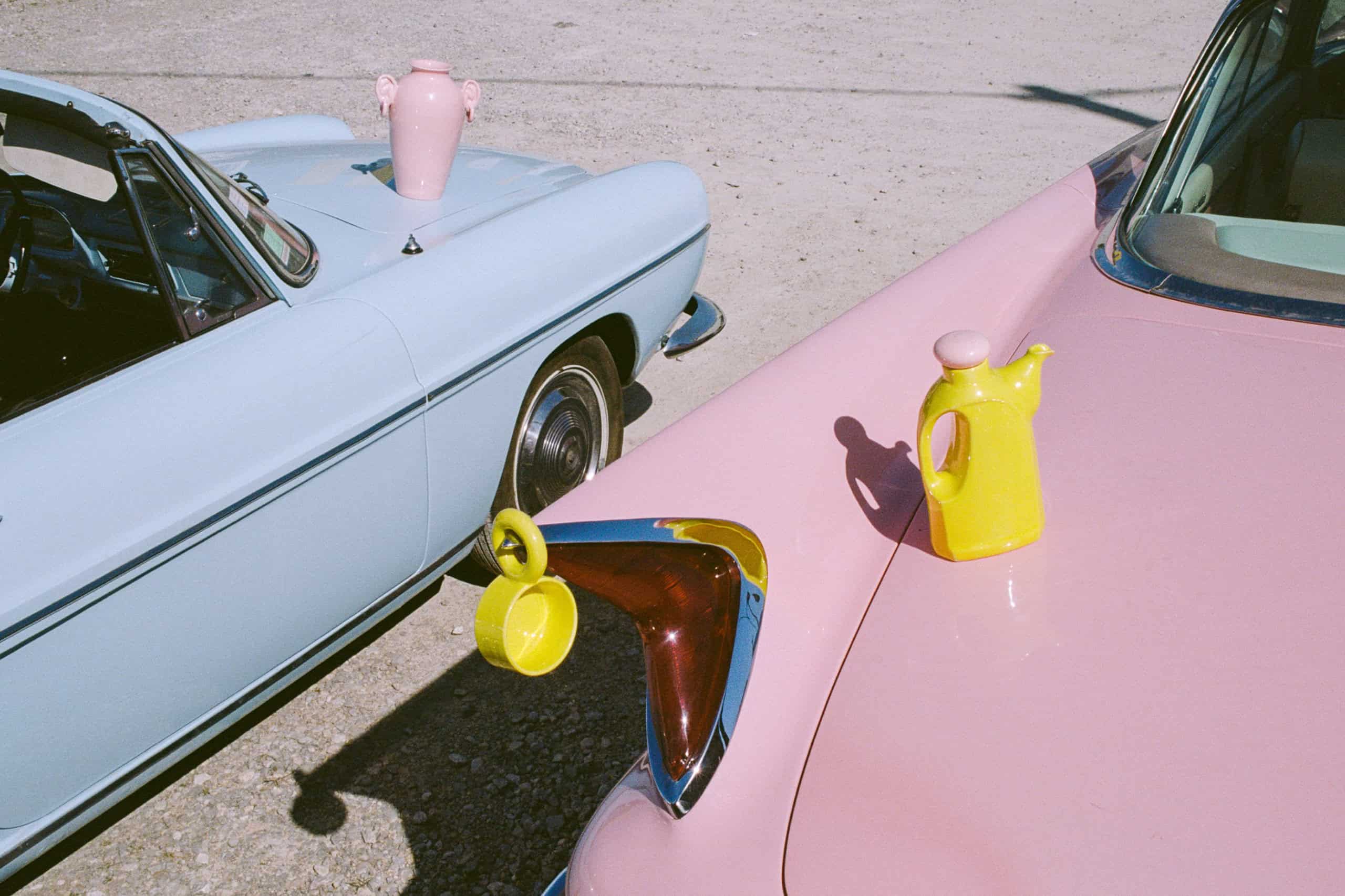 Two vintage pastel cars with ceramic objects placed on their hoods: a pink amphora, a yellow teapot, and a yellow cup. Artwork by Lola Mayeras, photographed outdoors in bright sunlight.