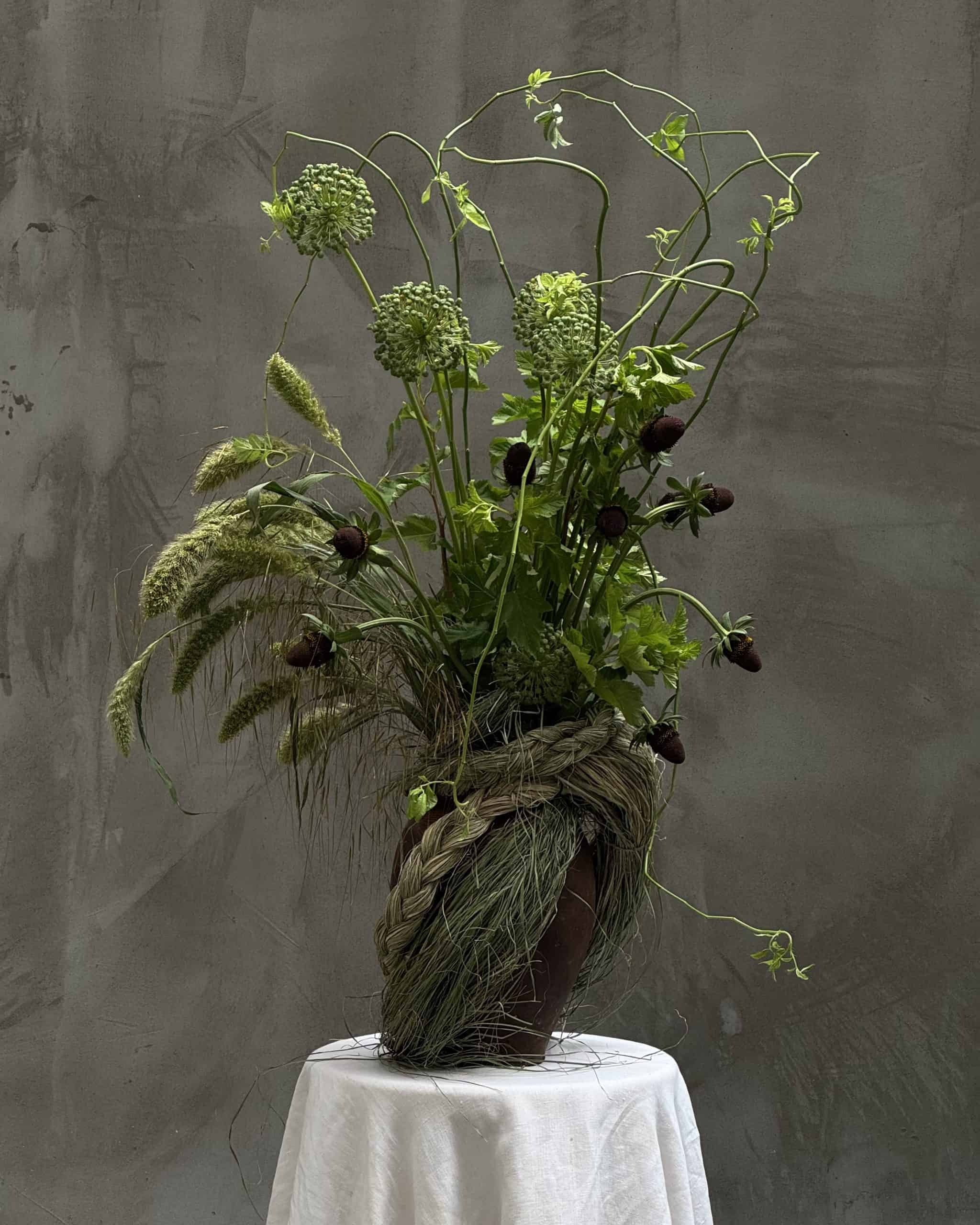 BLOOM CHRONICLES Green and textural floral sculpture in a twisting arrangement on a round white-draped table, by Mutabor.