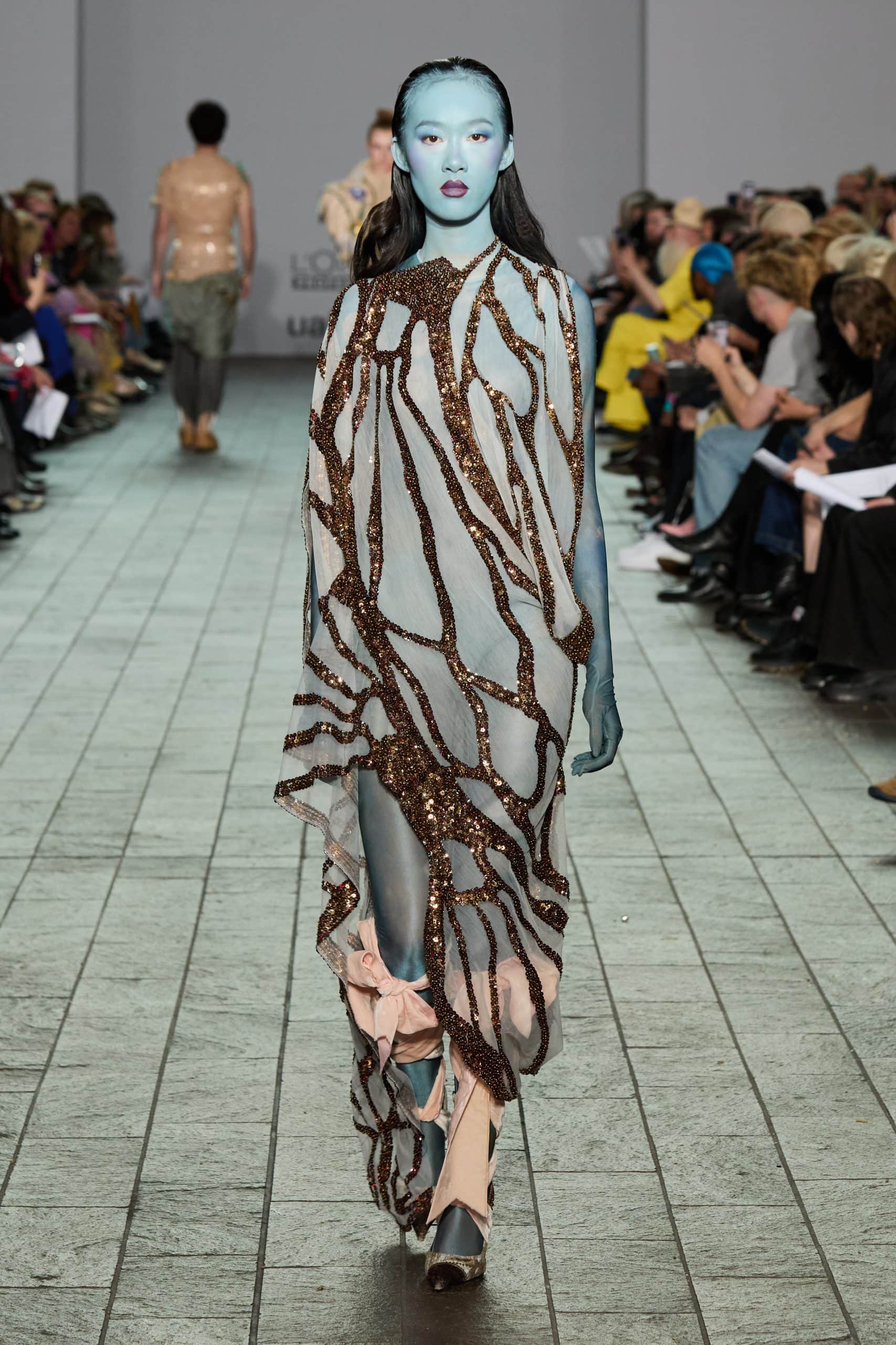 Runway model with painted blue face wearing a draped, branch-like textured gown . Courtesy of the designer