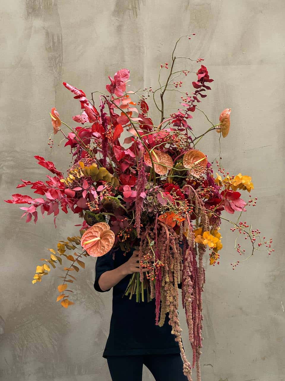 Person holding a large, overflowing floral arrangement of bright reds, oranges, and yellows against a concrete wall.BLOOM CHRONICLES
