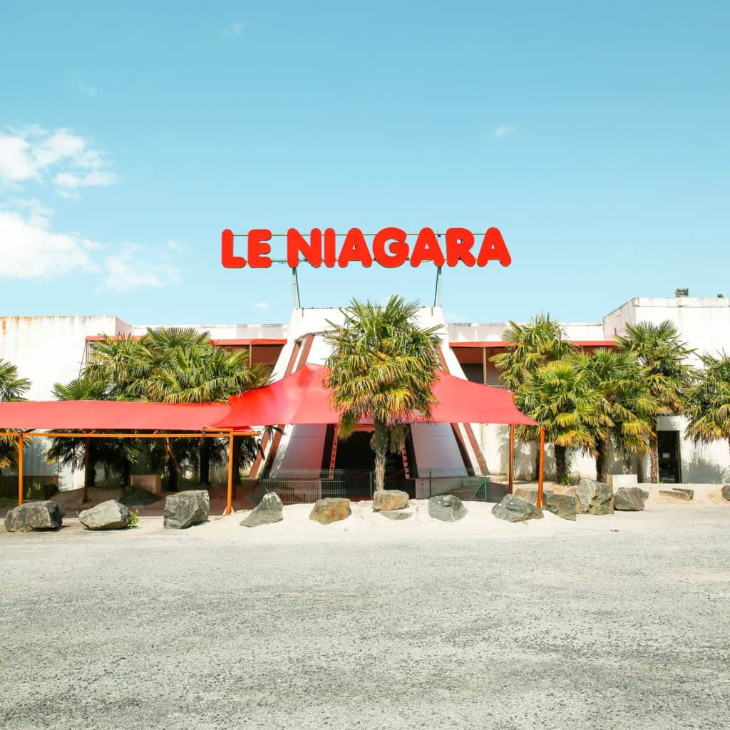Facade of Le Niagara nightclub with red awnings and palm trees, photographed in daylight. AFTER PARTY 