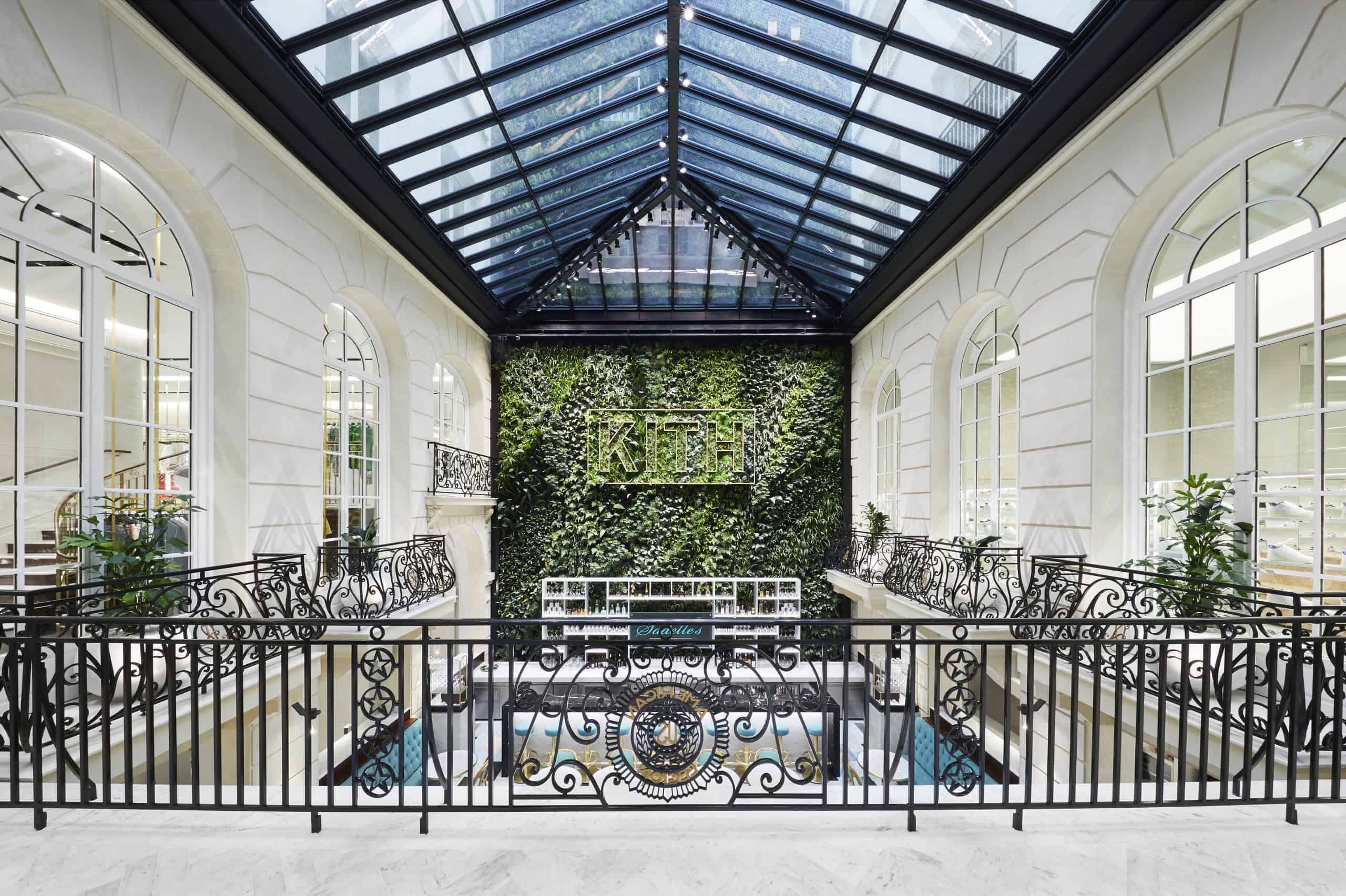 Interior courtyard of KITH Paris with a glass roof, white arches, wrought-iron details, and a green wall featuring the KITH logo.