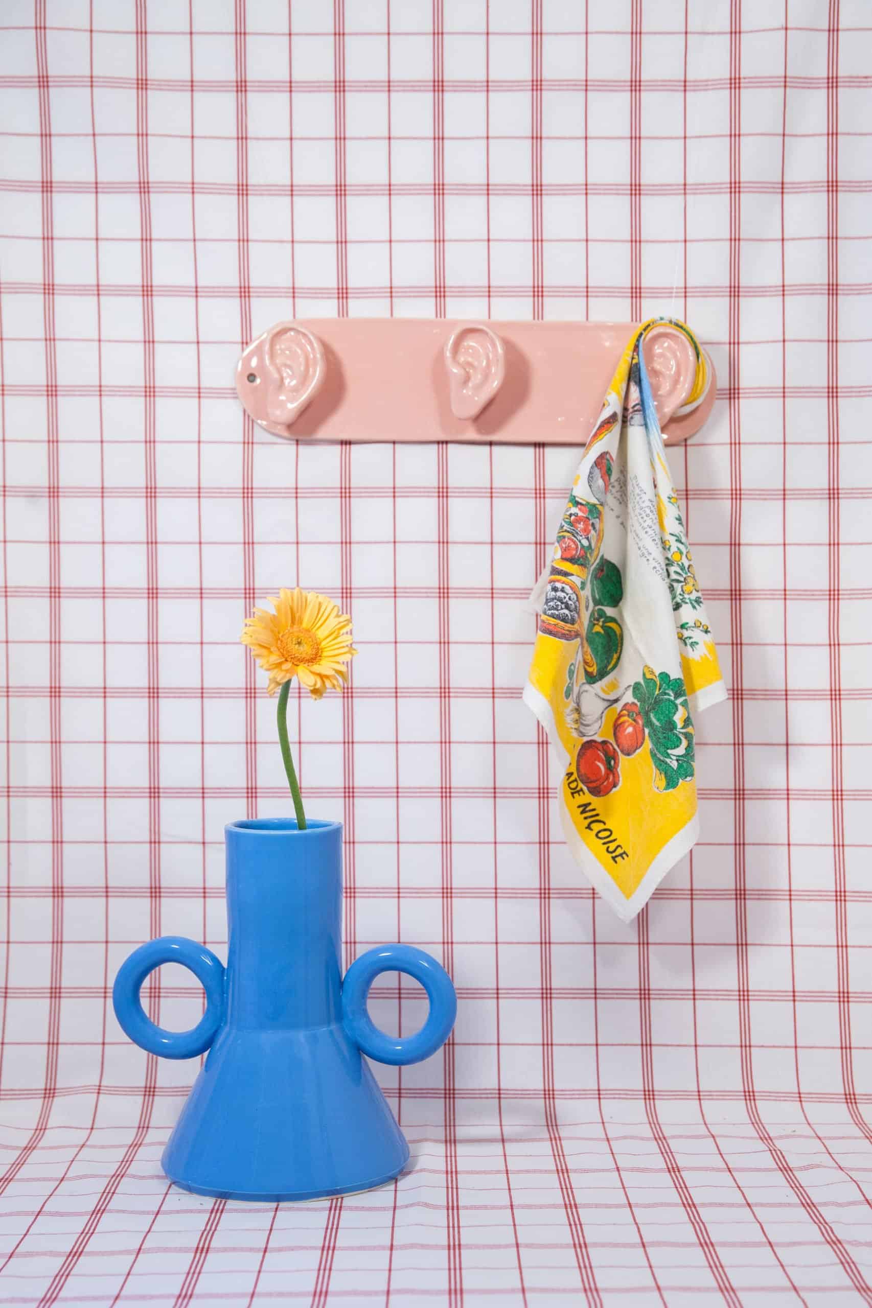 A whimsical ceramic scene by Lola Mayeras: a blue two-handled vase with a yellow flower sits below a pink wall hook shaped like three ears, holding a colorful dish towel. Set against a red grid fabric backdrop.