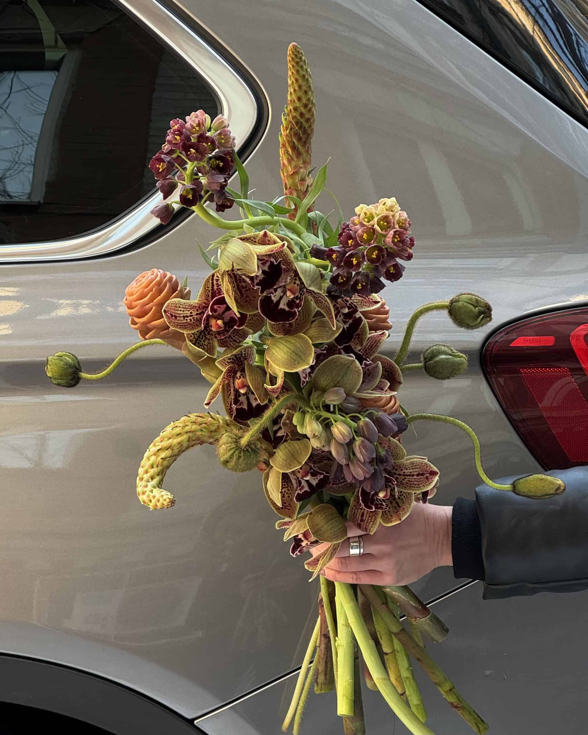 BLOOM CHRONICLES: Hand holding an asymmetrical bouquet with muted tones in front of a silver car, captured by Mutabor in Kyiv.