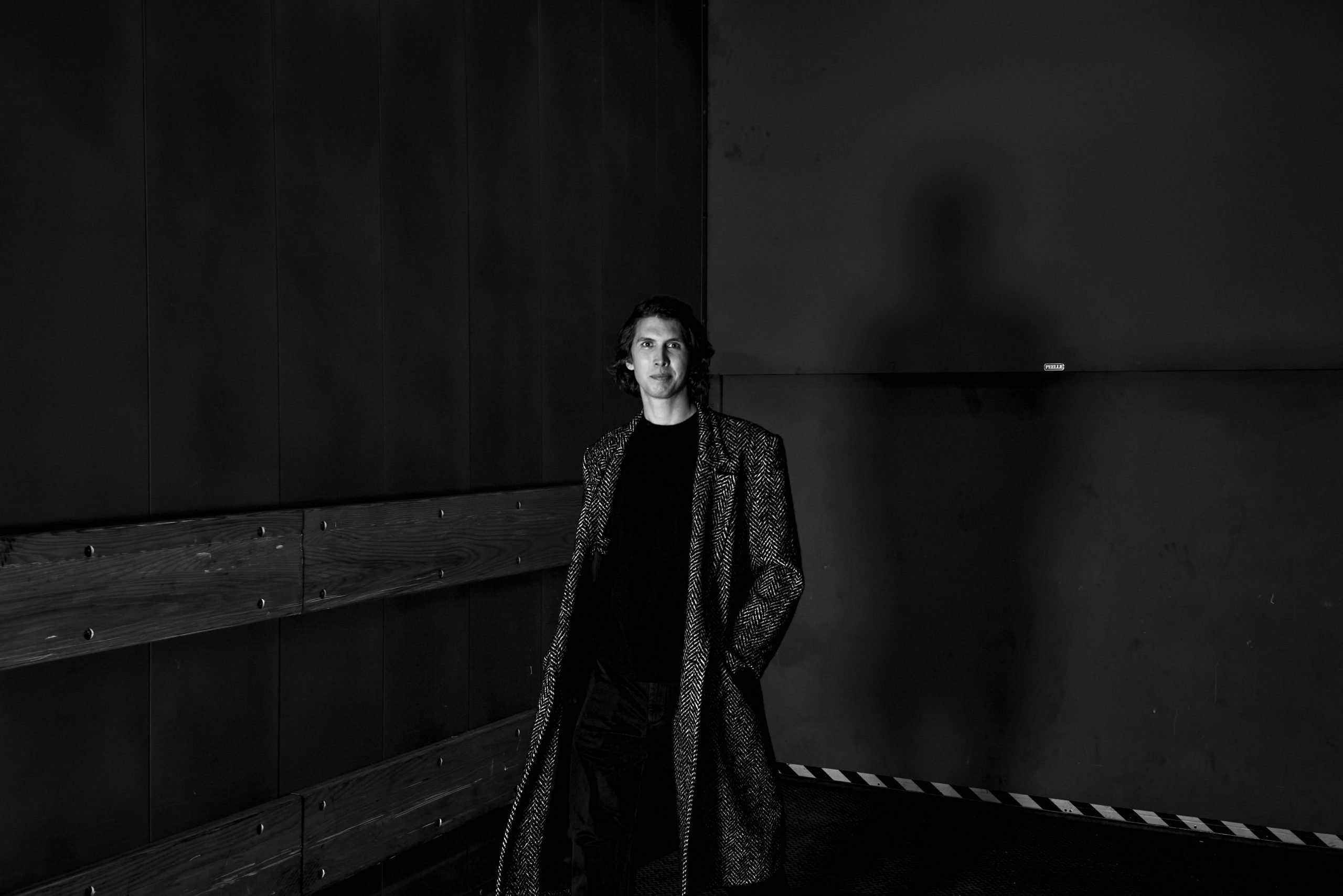 Andrew Boszhardt in a long coat, standing in a dimly lit industrial hallway, casting a shadow behind him. Photo by Van Strahl