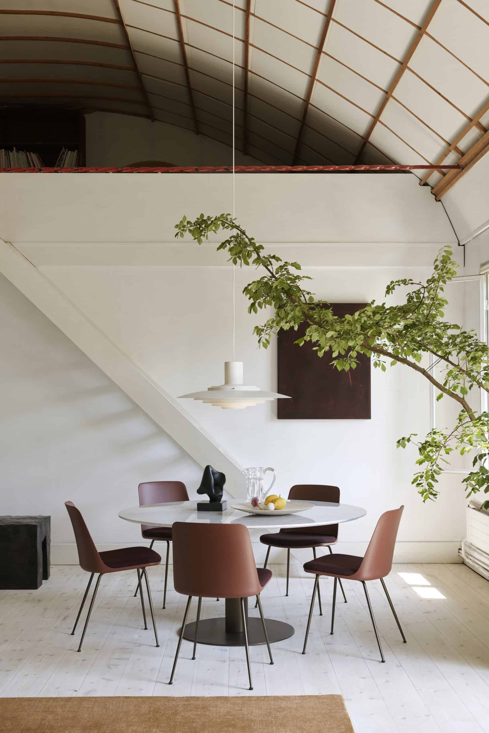 The P376 Pendant by &Tradition — a sculptural form suspended above a round dining table with burgundy chairs, framed by angled beams and lush indoor greenery.