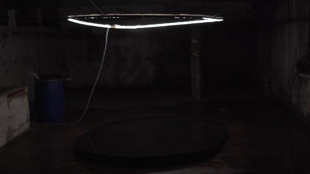 Installation view of Juju Bento's artwork "Rundemanen" from 2022. Tiny drops of water fall off of an illuminated steel circle, suspended in air.