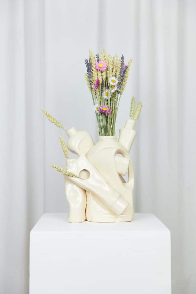 A cream-colored sculptural vase resembling stacked detergent bottles, filled with dried flowers, designed by Lola Mayeras.