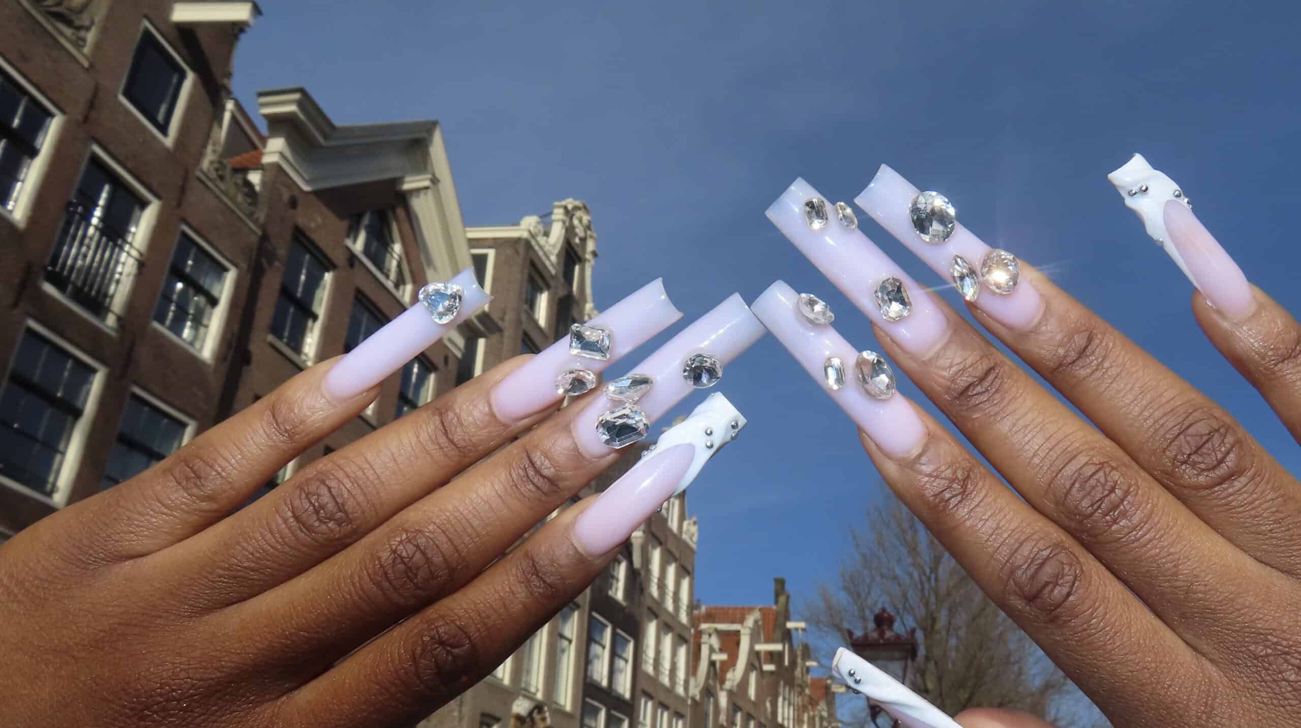 Close-up of hands with long white rhinestone-studded fake nails, held up in front of a brick building.