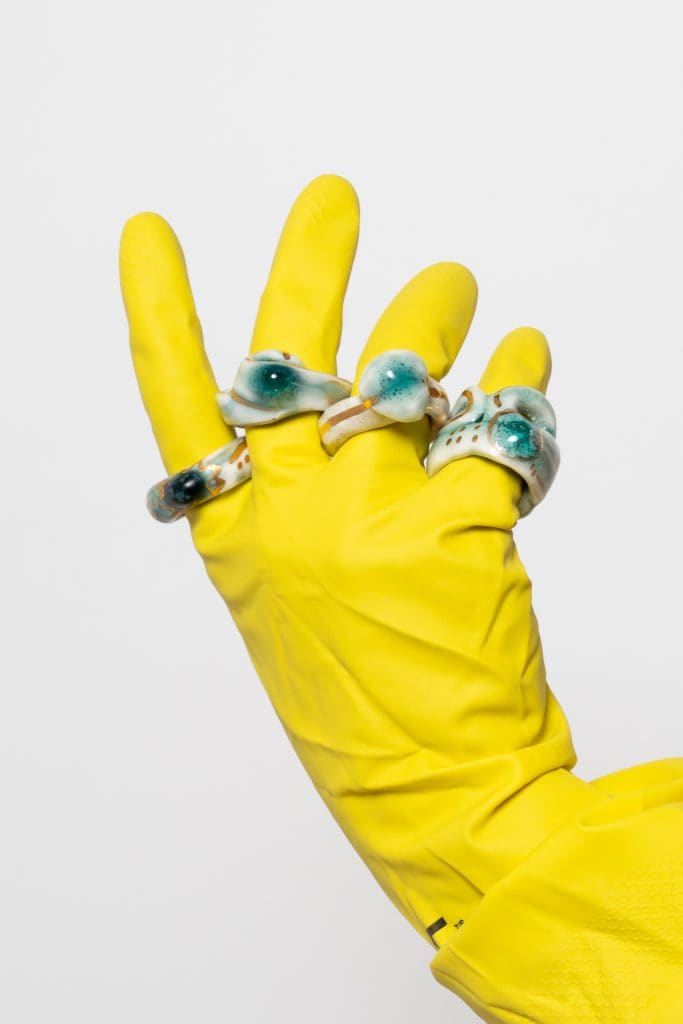 Yellow-gloved hand wearing multiple ceramic rings, raised in mid-motion. JORDANE SOMVILLE