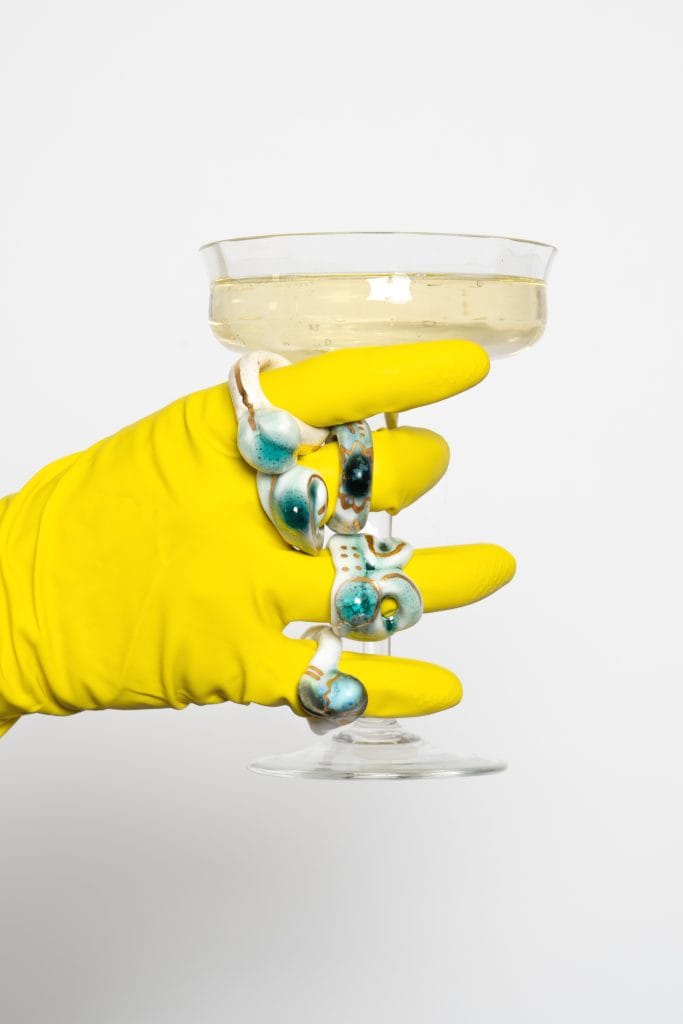 Champagne glass held by a yellow-gloved hand adorned with glazed rings.