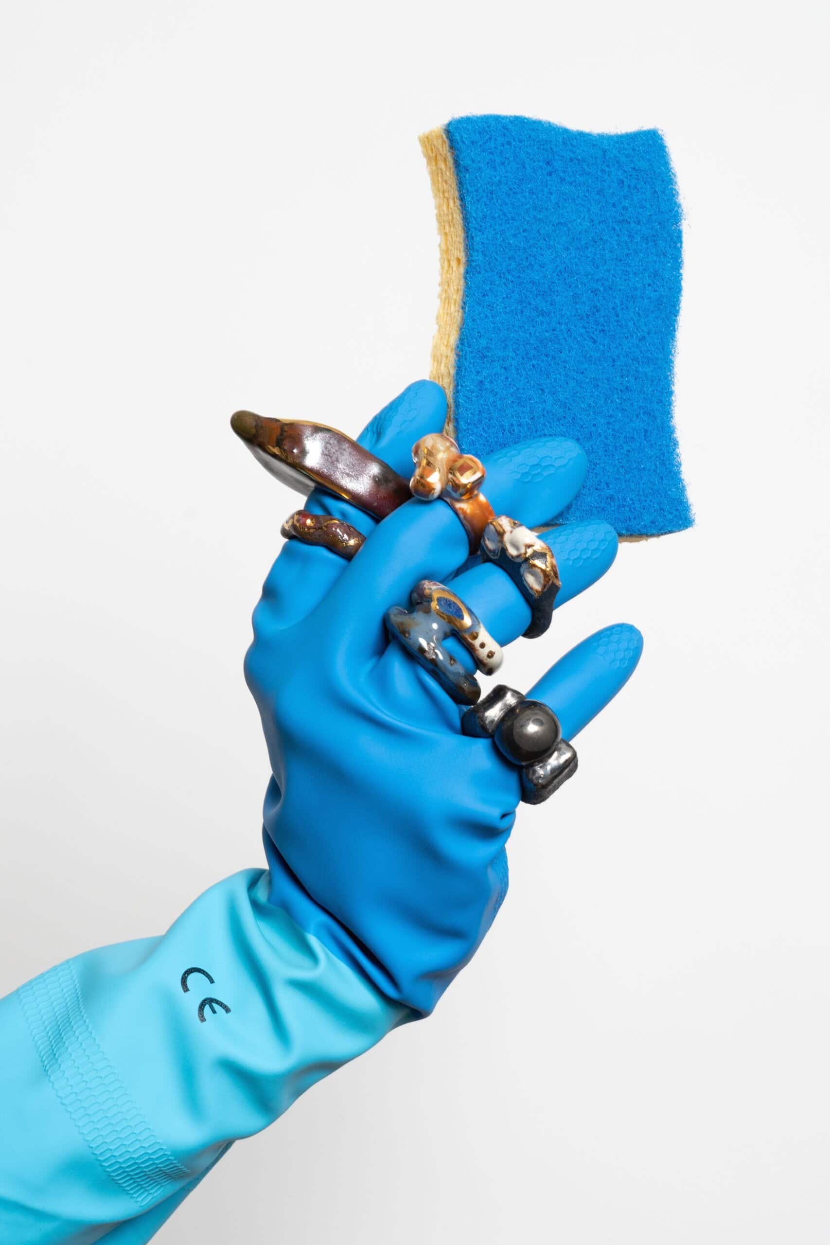 A gloved hand adorned with ceramic rings holds a blue cleaning sponge against a white backdrop.
