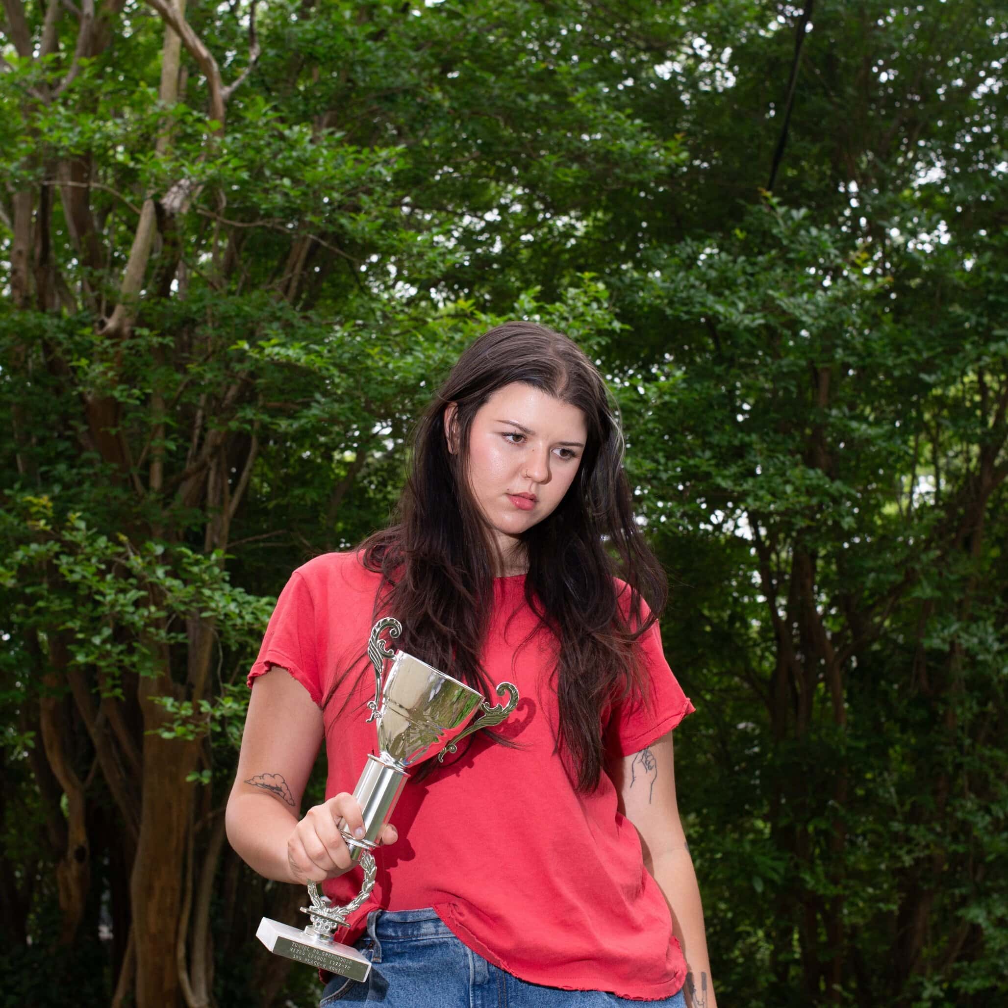 Eliza McLamb stands outdoors holding a handmade trophy, wearing a red t-shirt, looking contemplative.
