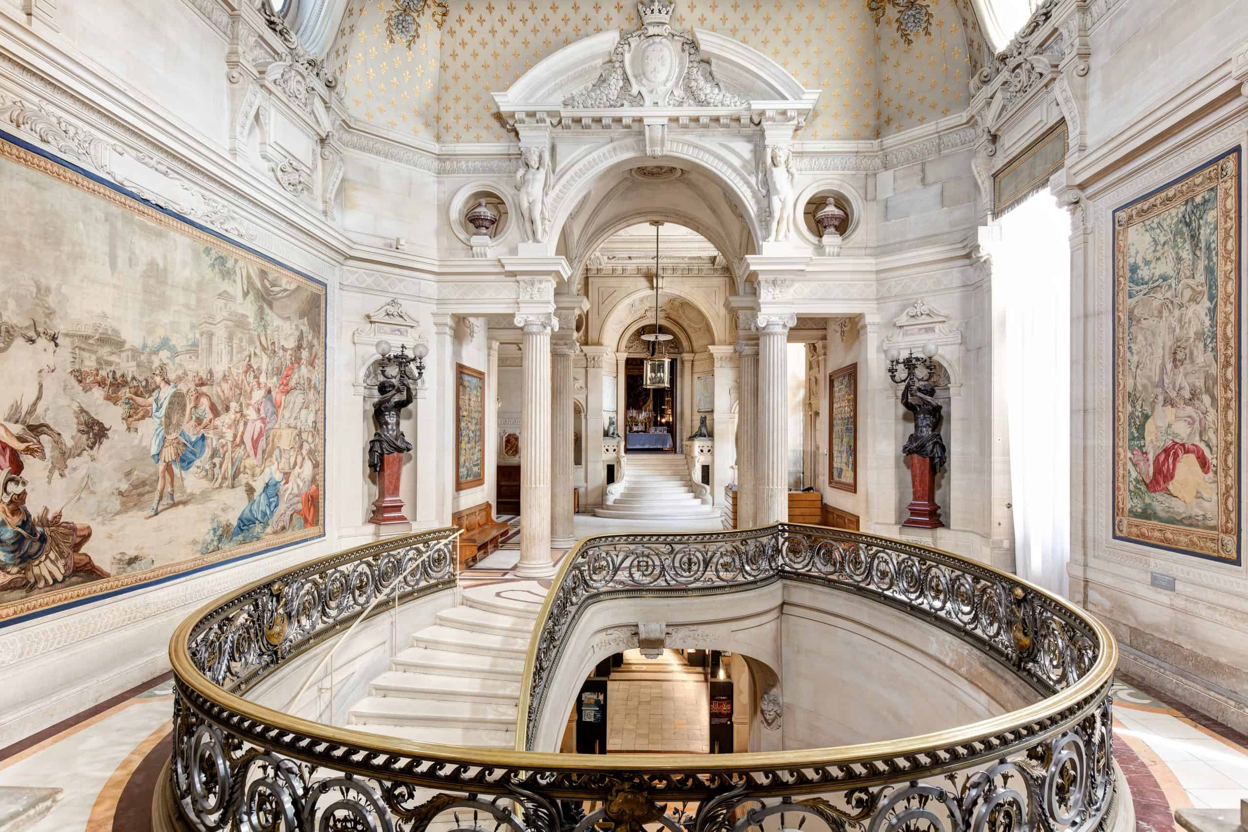 Interior view of Château de Chantilly’s grand staircase, with ornate railings, classical statues, and rich wall tapestries. Courtesy of Domaine de Chantilly.