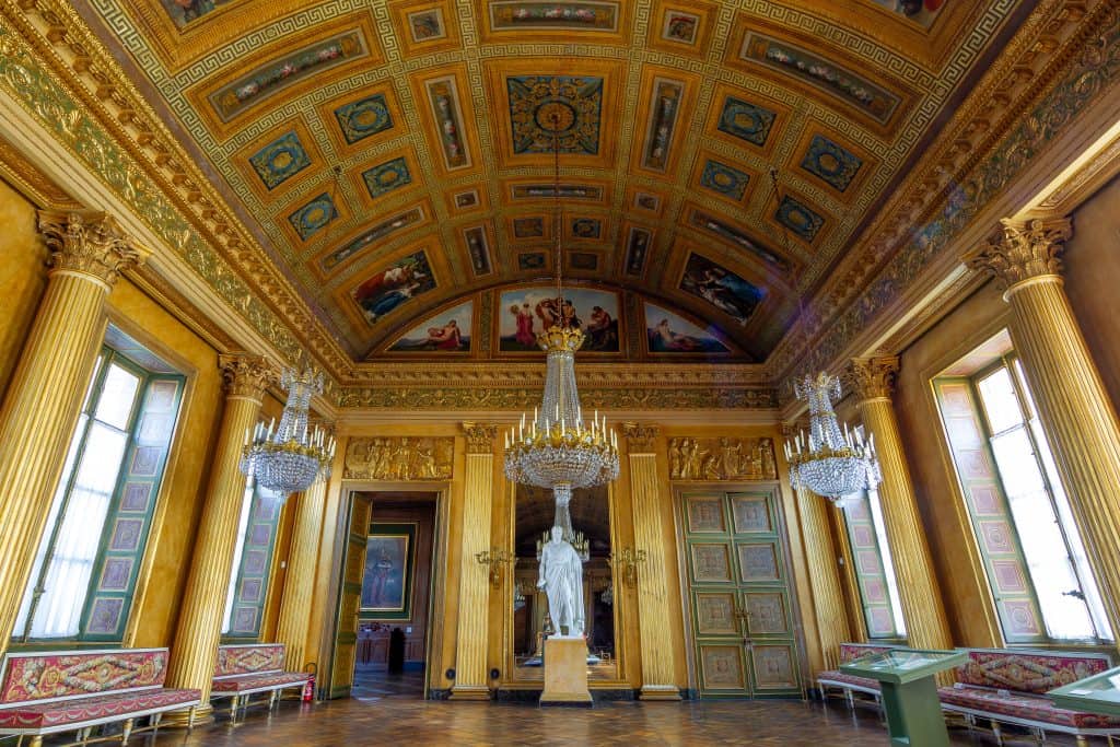 Ballroom of Compiègne with golden ceiling panels, grand columns, and ornate chandeliers. Courtesy of Centre des monuments nationaux.