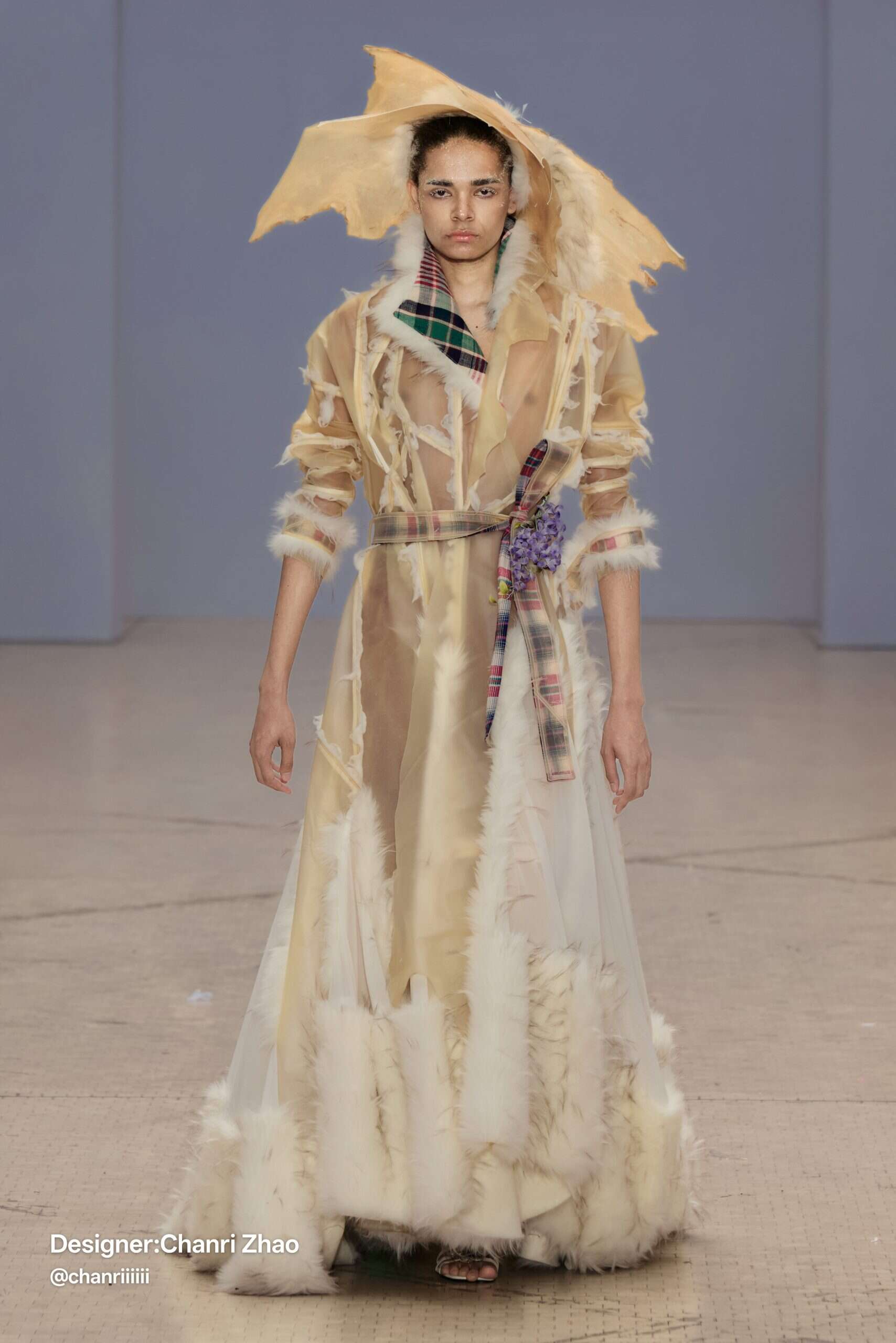 Model wears a translucent floor-length coat trimmed with faux fur and tartan lining, topped with an abstract sculptural hat—by Chanri Zhao. Courtesy of designer Chanri Zhao.