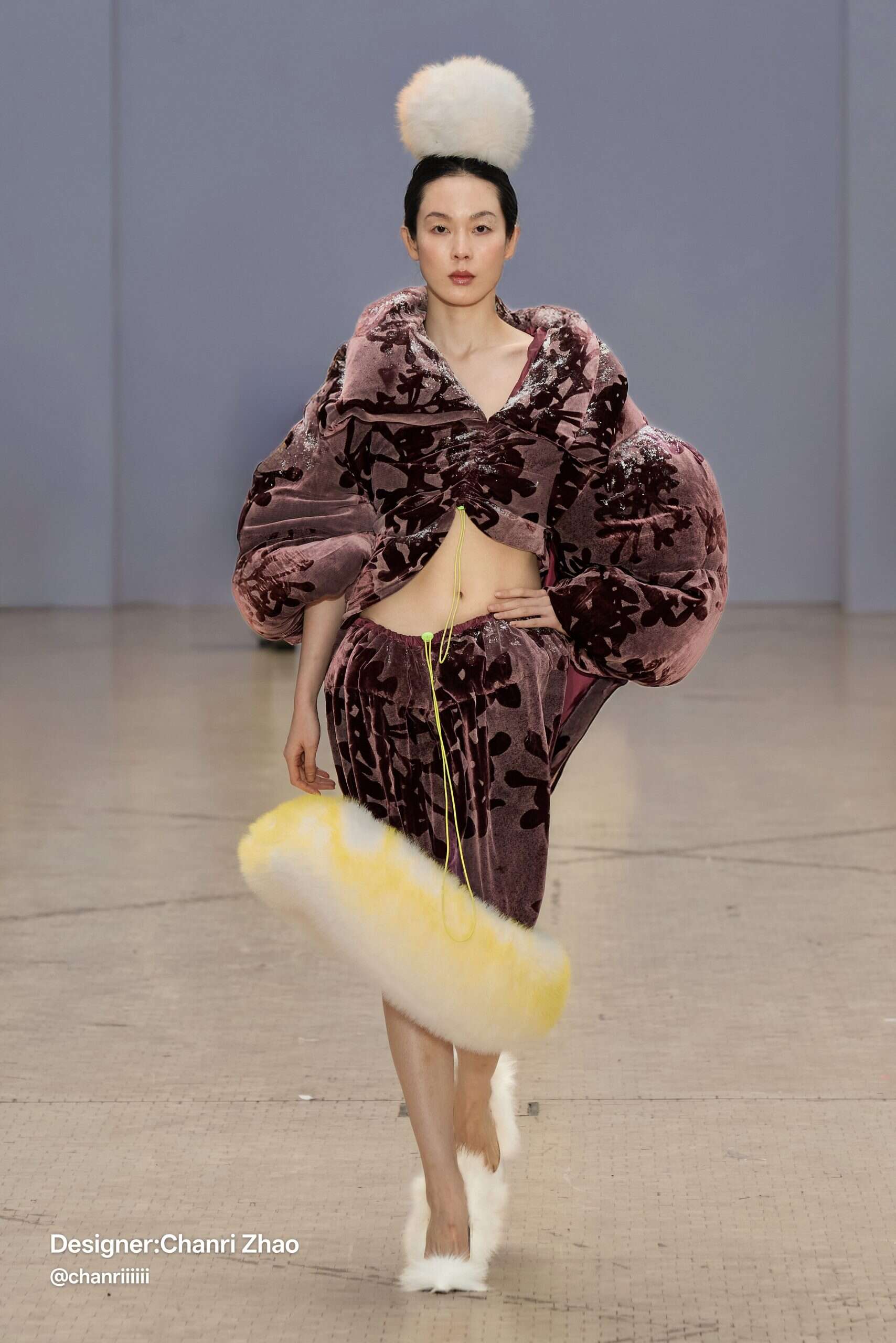 Model walks in a purple velvet ensemble with floral motifs, voluminous sleeves, fur-trimmed skirt, and a pompom headpiece—by Chanri Zhao. Courtesy of designer Chanri Zhao.
