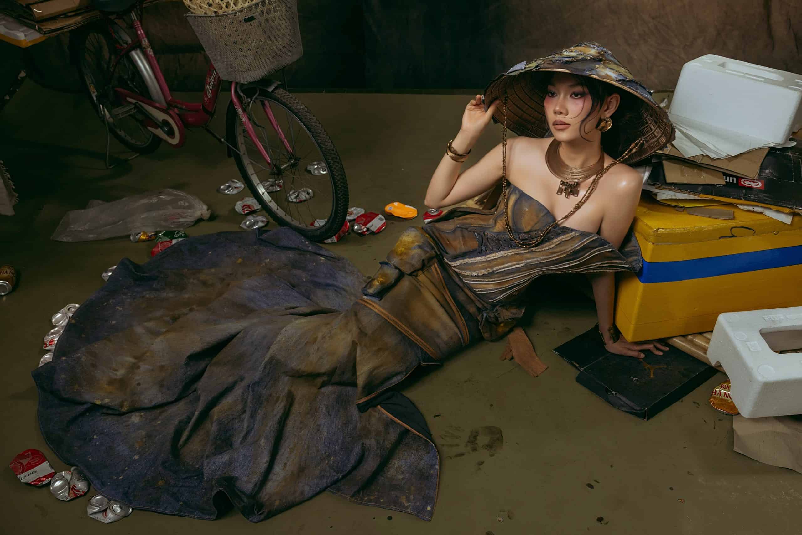 Model wearing rust-dyed gown lying on the ground surrounded by scrap objects. IAM – SS’25, resilience woven into rust and denim. Courtesy of Célianne Nhatlam.