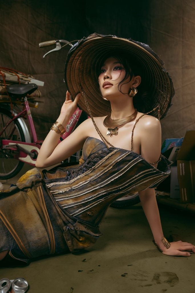 Model wearing scrap-textured gown and large hat, seated on the floor.