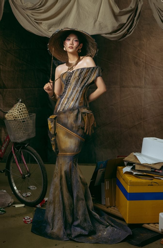 Model in scrap-inspired gown standing beside bicycle and boxes. IAM – SS’25, resilience woven into rust and denim. Courtesy of Célianne Nhatlam.