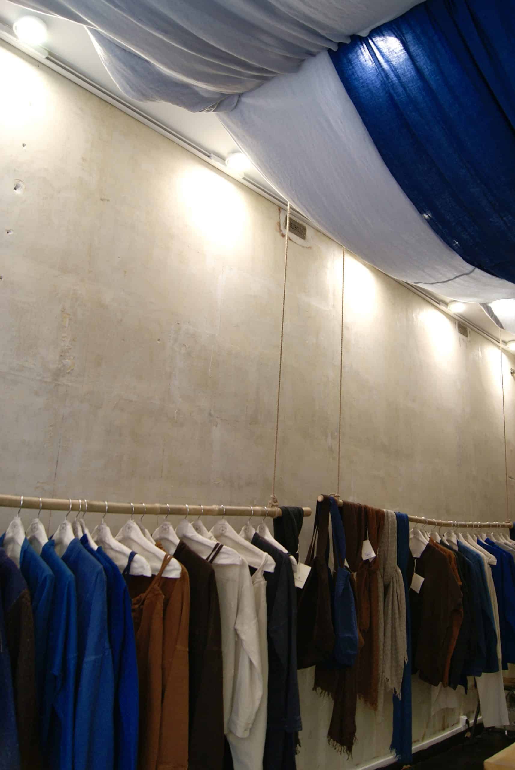Clothing racks with blue and neutral garments beneath woad-dyed fabric ceiling at Bleu de Cocagne Paris.