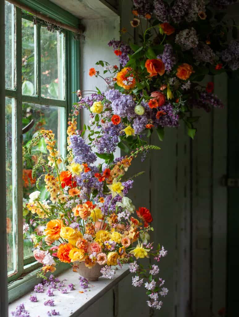 Tall, airy flower arrangement in purples and greens, placed in a dark vase in front of a faded pink cloth.