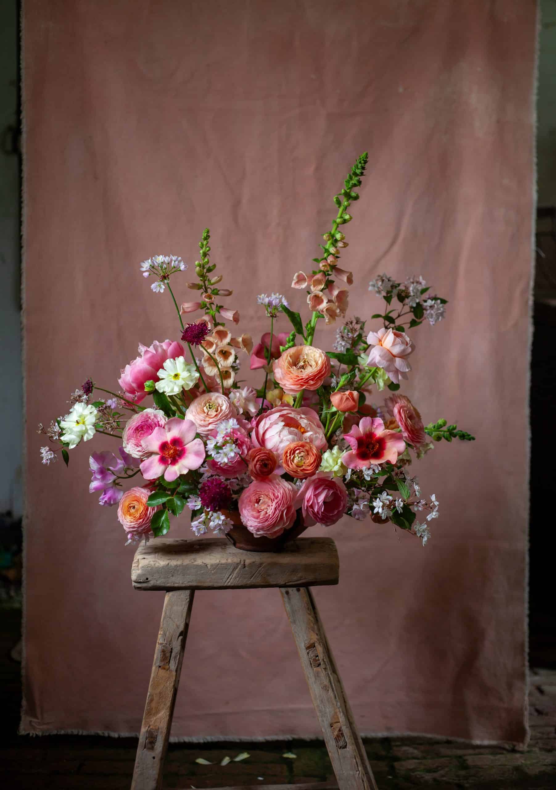 Vase of lush pink and coral arranged asymmetrically on a rustic stool, with a soft rose-colored backdrop.