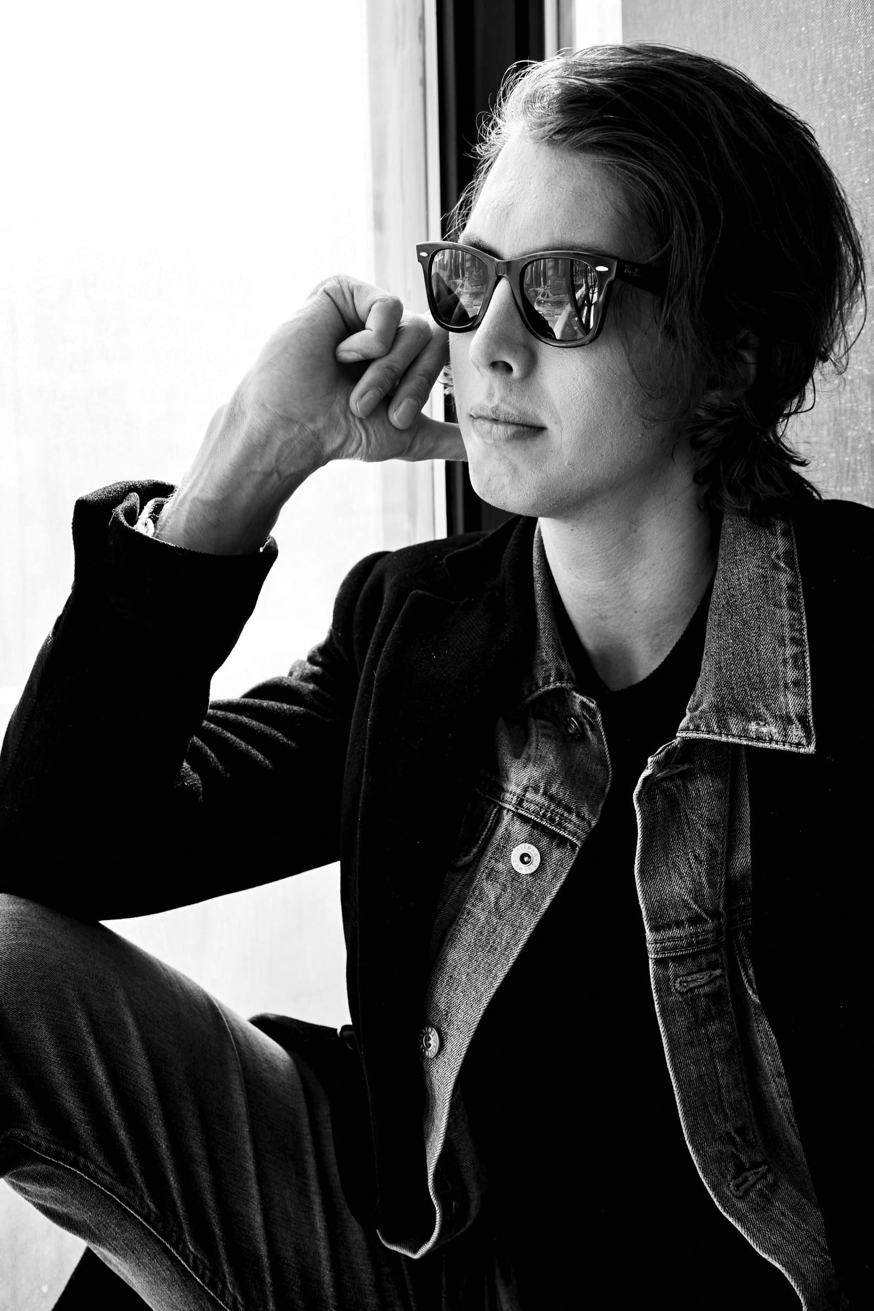 sitting by a window, wearing sunglasses and a layered denim and blazer look, gazing into the distance.