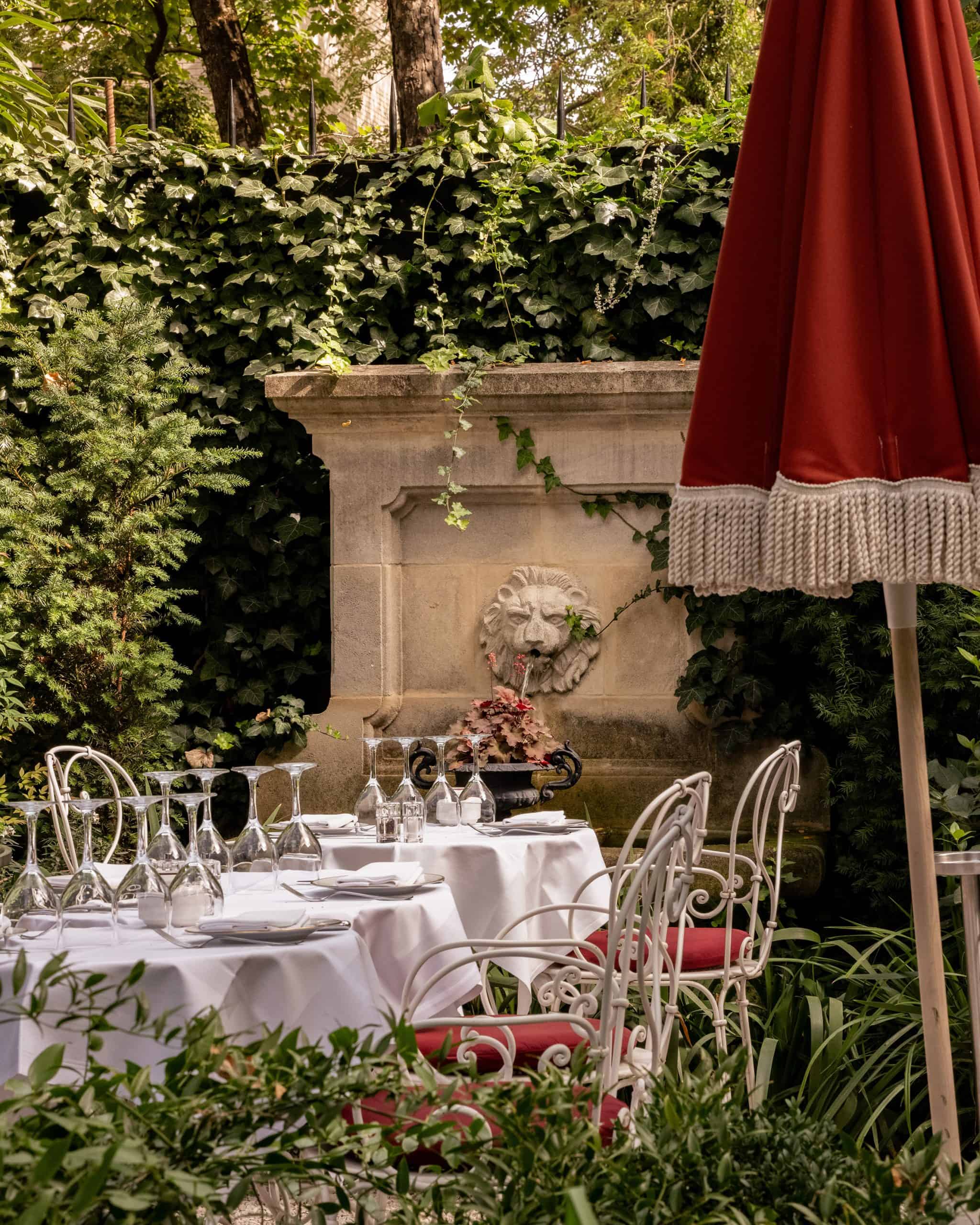 A secluded garden dining area at Hôtel Particulier Montmartre, with white tablecloths, red chairs, and a lion-head fountain.
