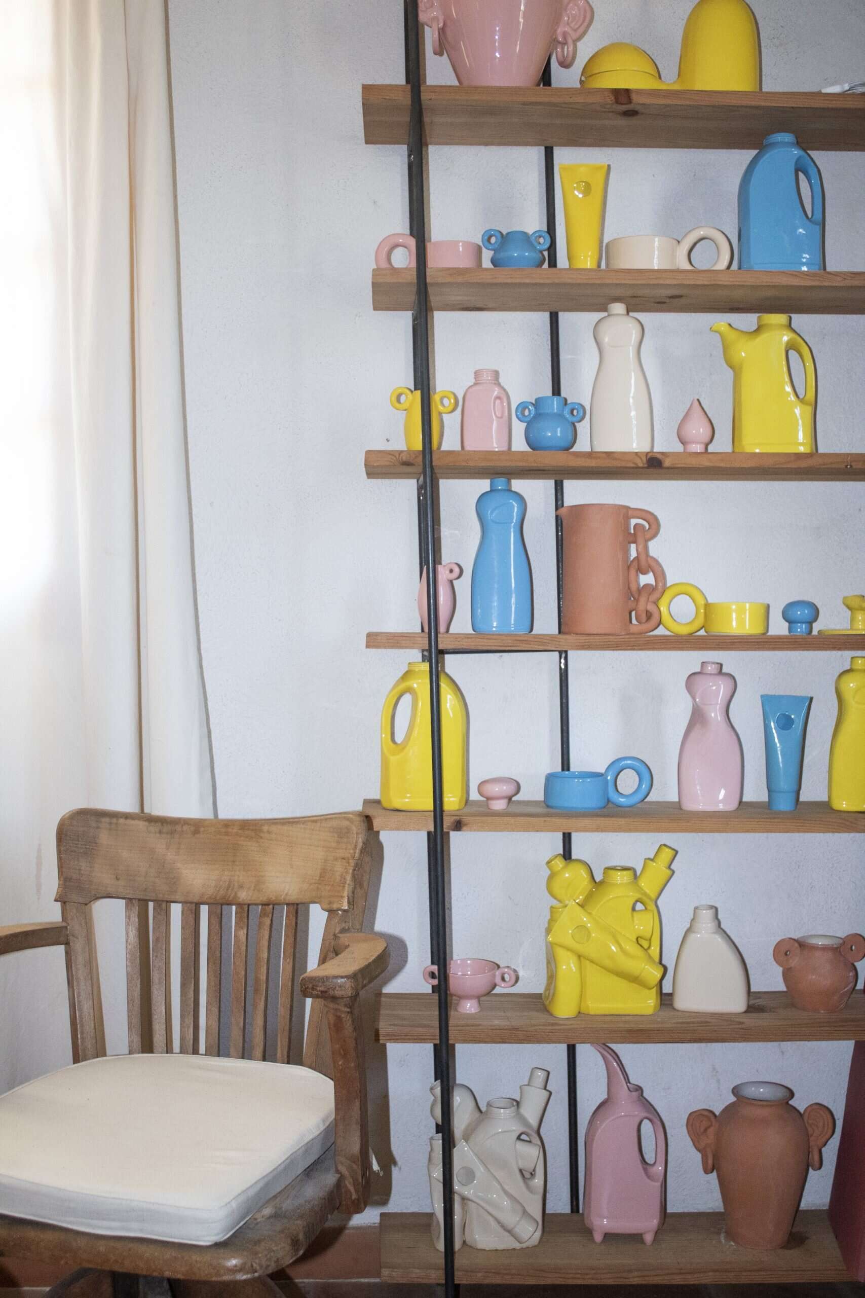 A wooden shelf in Lola Mayeras’ studio filled with colorful ceramic objects in yellow, pink, blue, and cream. The sculptural pieces include vases, jugs, and surreal forms. A rustic wooden chair sits beside the display.