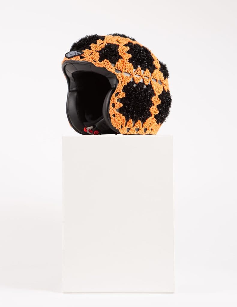  Black boxing helmet wrapped in orange granny-square crochet by Sophie Inard, displayed on a white pedestal.