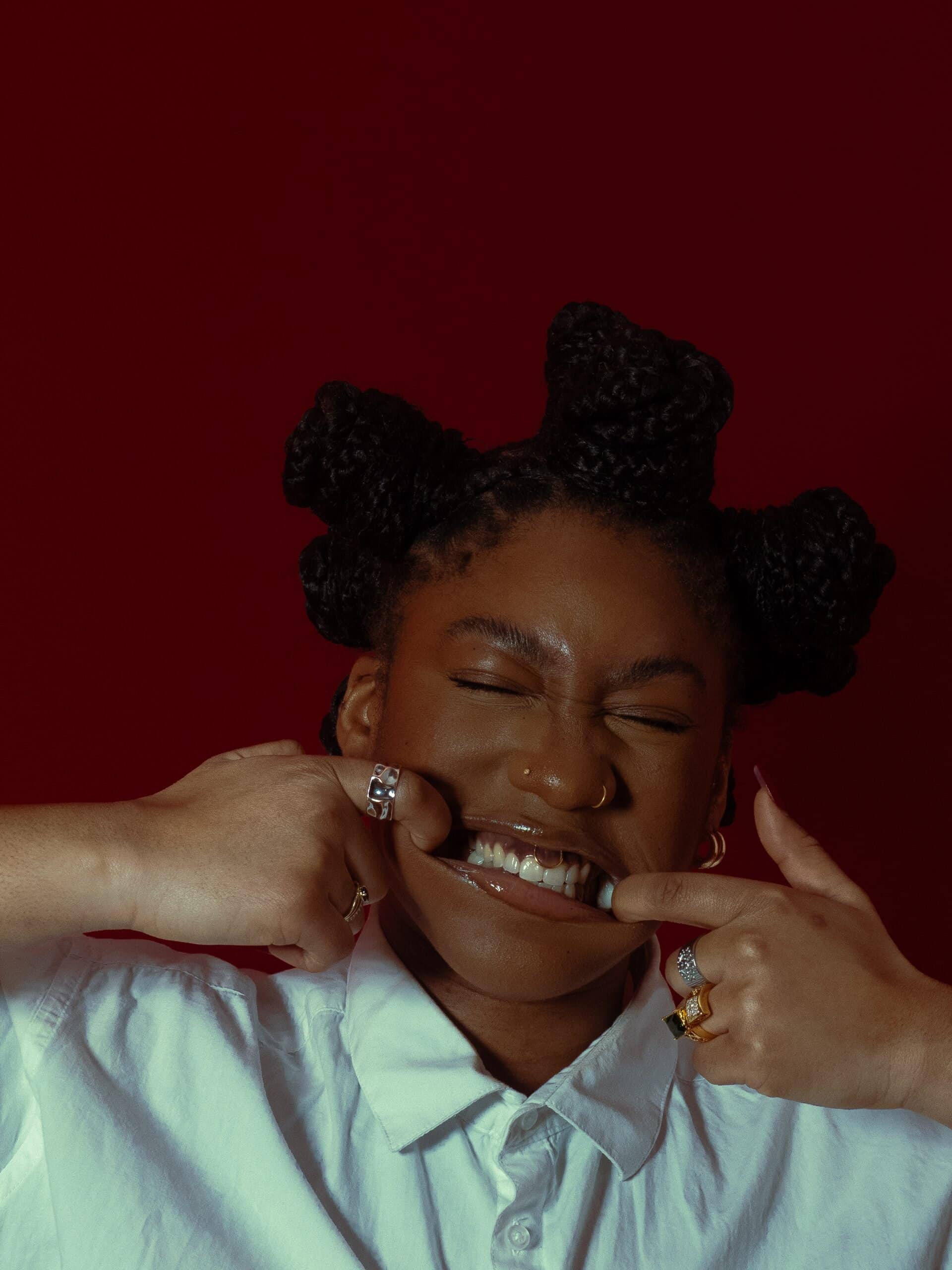 Model Jeanne Saindou makes a playful face, stretching her smile with both hands, wearing a white shirt and statement rings—photographed by Kloé Camier.  For undressed to impress 