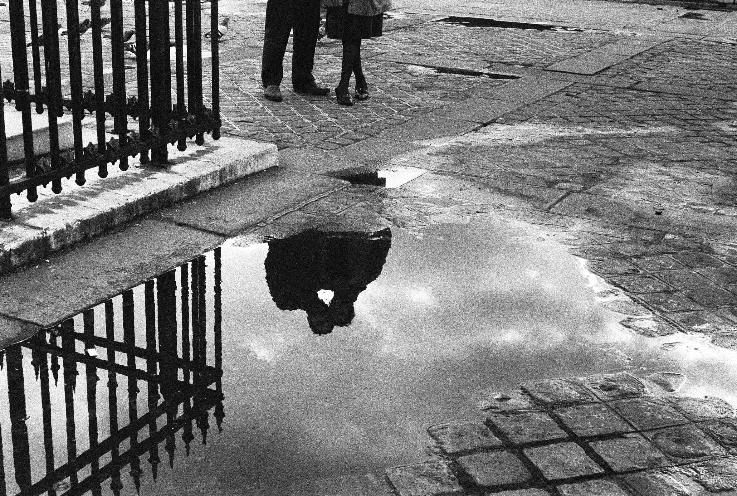 Black-and-white photograph of two people standing near an iron gate, their lower bodies visible, with their reflection captured in a puddle on a cobblestone street. Photo by Nicole Lala, courtesy of her daughter Delphine.