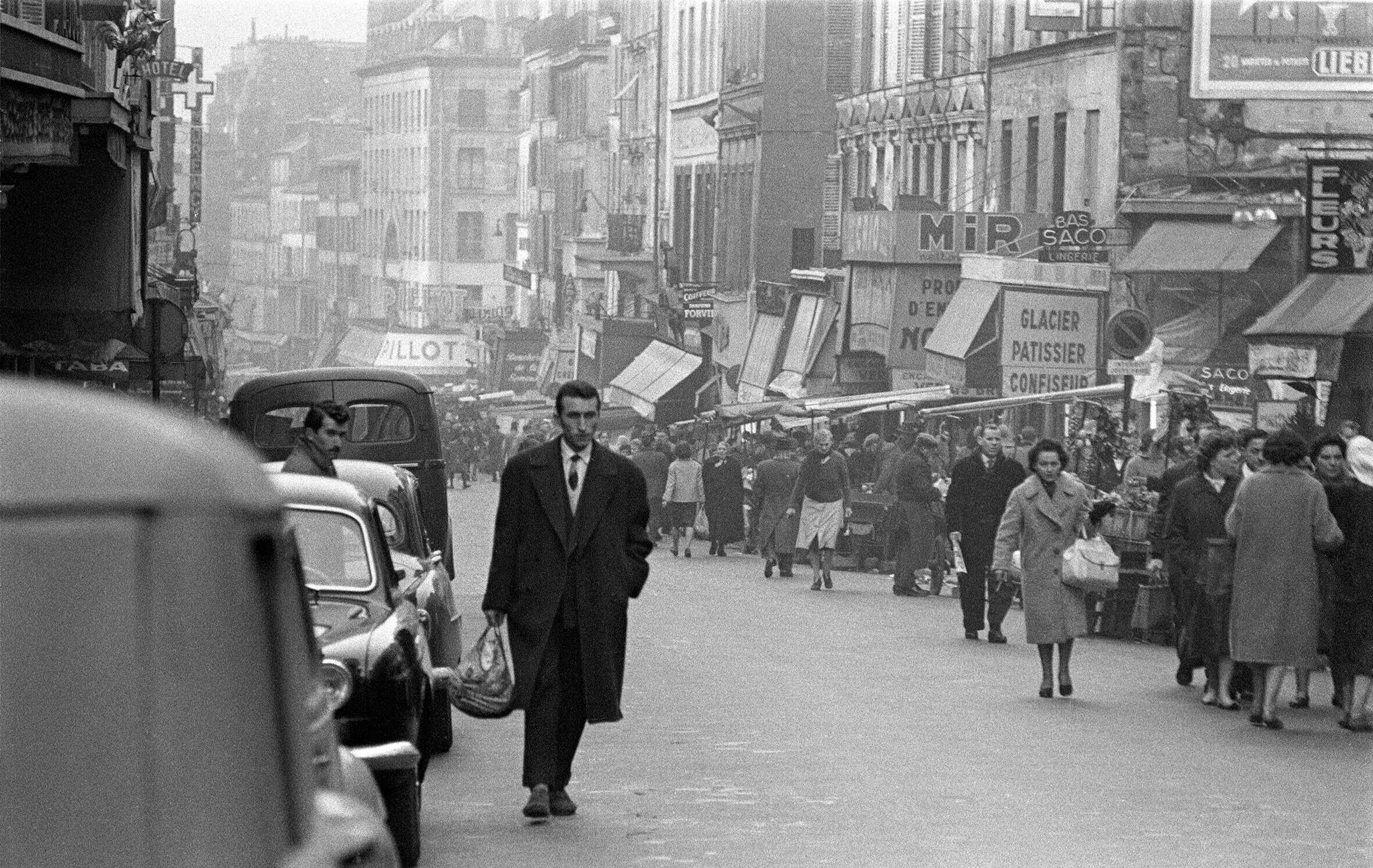 Black-and-white street scene in Paris showing a crowded avenue filled with pedestrians, market stalls, vintage cars, and storefront signs. Photo by Nicole Lala, courtesy of her daughter Delphine.