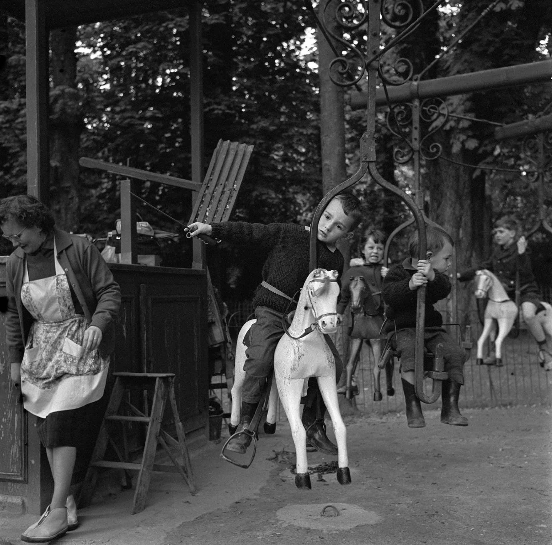 
Black-and-white photo of children riding a vintage carousel with carved wooden horses, while a woman stands nearby. Photo by Nicole Lala, courtesy of her daughter Delphine.