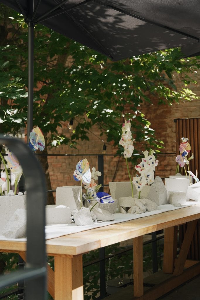 Outdoor display of hand-painted flowers hanging and standing, reimagining bridal bouquets as art in a lush garden setting.