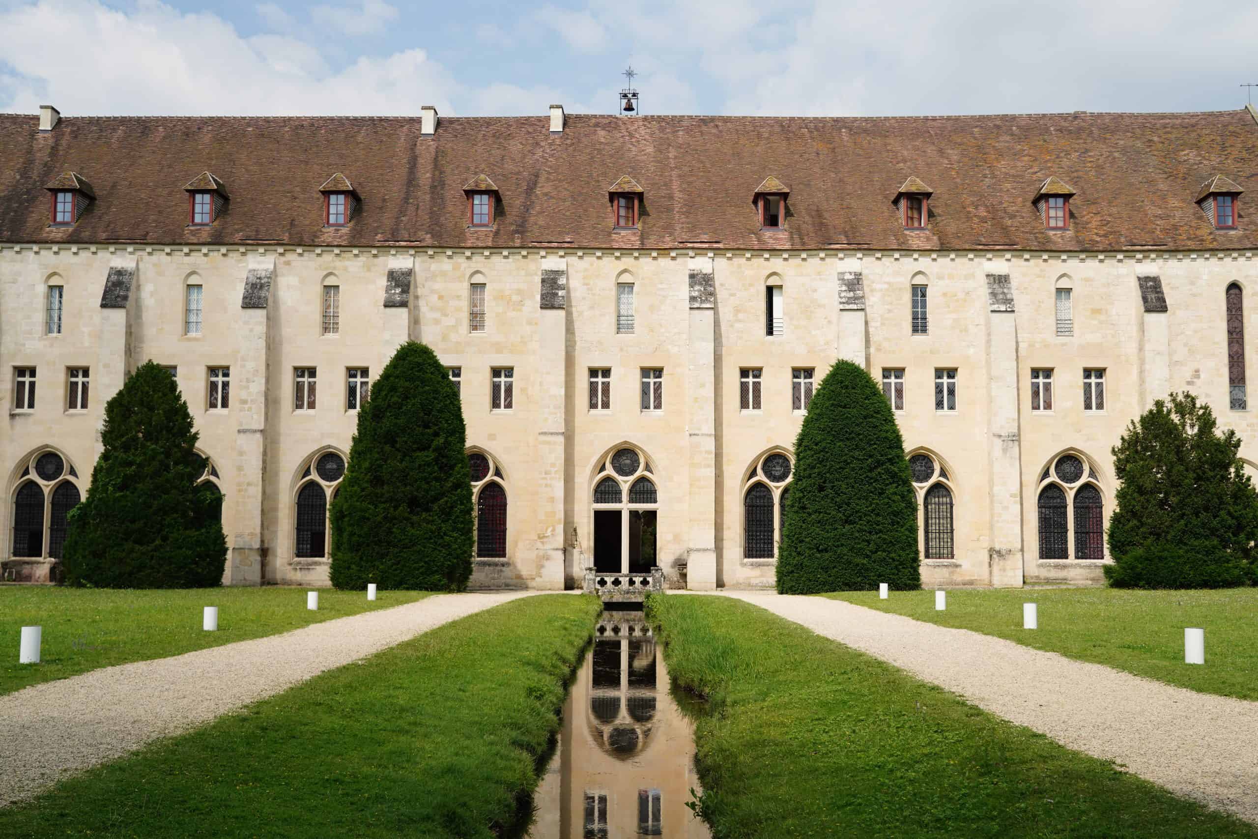 Front view of Royaumont Abbey’s monk building with trimmed trees, a central pond, and a medieval stone façade. Courtesy of Royaumont Foundation.