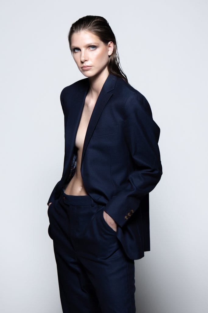 Cornelia in deep navy suit, bare chest, powerful stance, minimalist styling.
