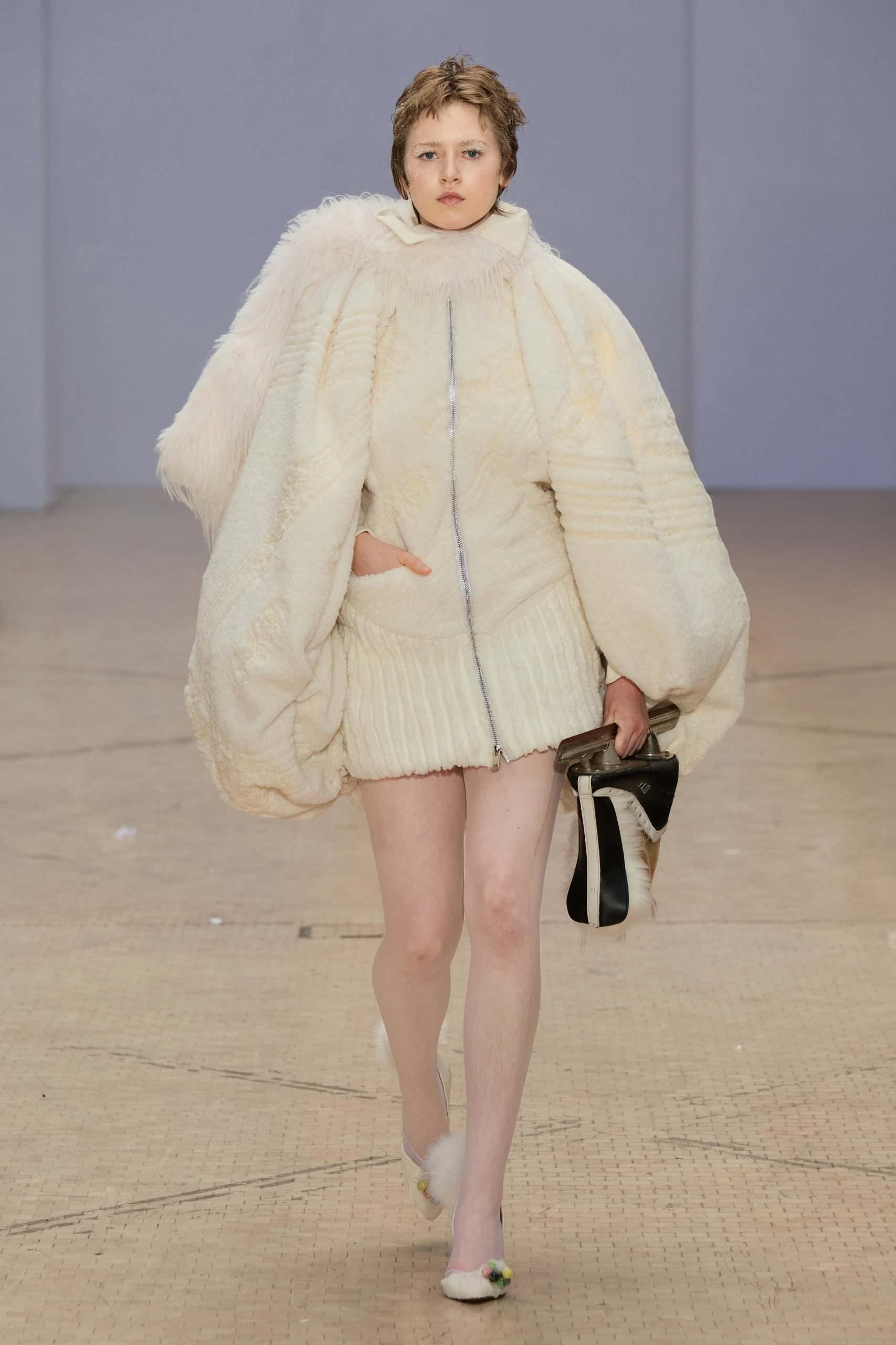 Model wears an oversized cream shearling cape with subtle patterns, ribbed mini-skirt, and soft slippers—designed by Chanri Zhao.
