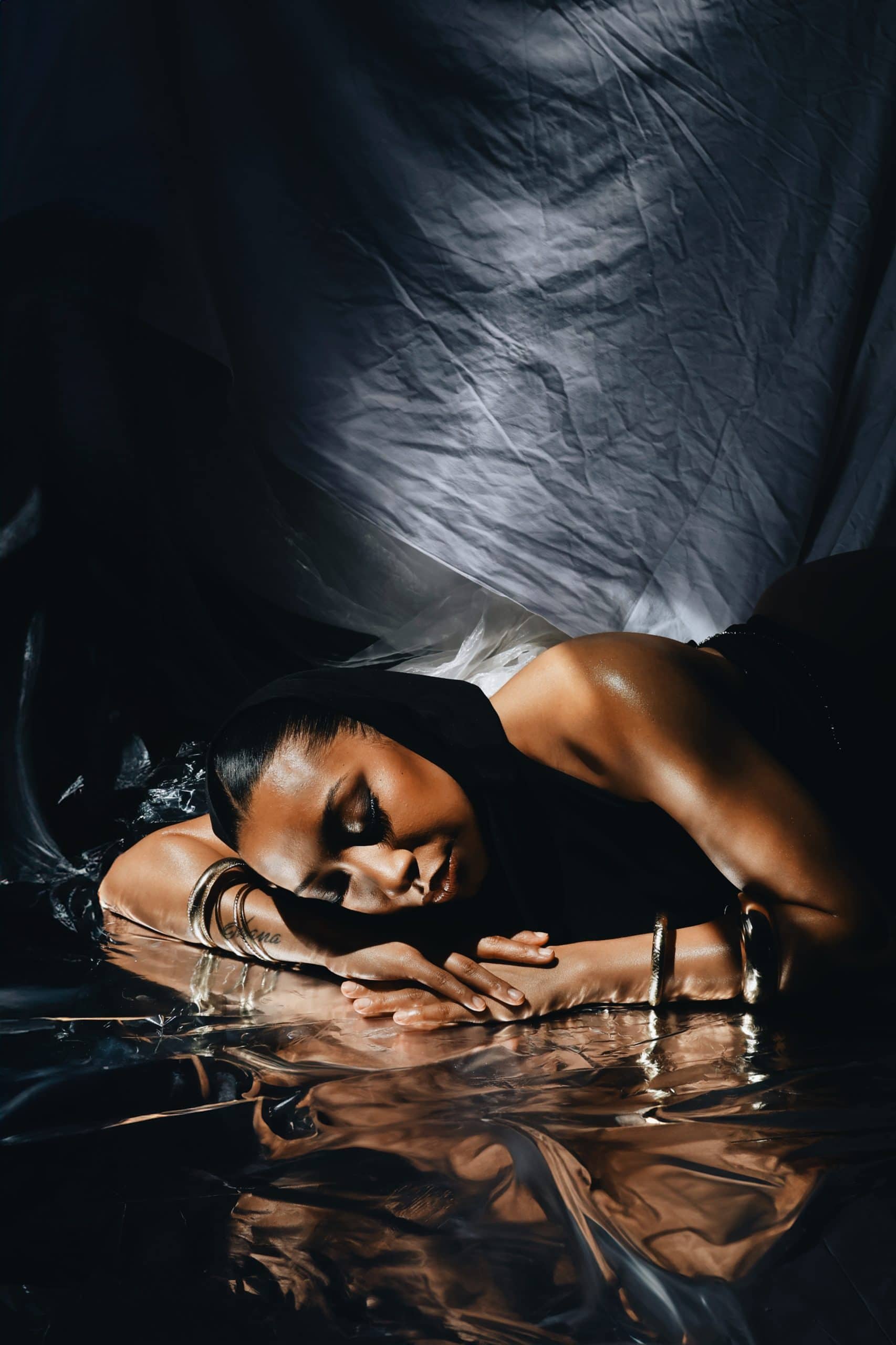 A female rapper lies in soft light, draped in black fabric, her reflection cast in metallic foil—evoking strength, vulnerability, and introspection. For undressed to impress 